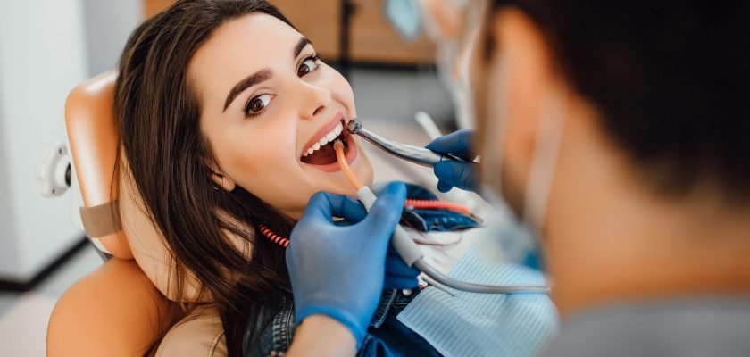 Young female patient visiting dentist office. Beautiful woman with healthy straight white teeth sitting at dental chair with open mouth during oral checkup while doctor working at teeth.